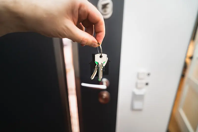 Schlüsselbund mit Haus-Anhänger in einer Hand, dahinter eine angelehnte Wohnungstür und eine helle Wand