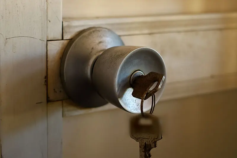 Ein Schlüssel mit Ring steckt im Schloss eines runden Metallknaufs an einer Holztür, darunter hängt ein zweiter Schlüssel