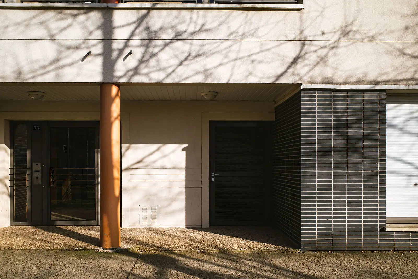 Eingangsbereich eines Mehrfamilienhauses mit Glastüren und Klingelanlage, davor der Schatten von Ästen im Streiflicht